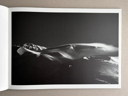 Open photobook spread showing a black-and-white landscape photograph of snow-covered dunes or ridges under a dark sky.