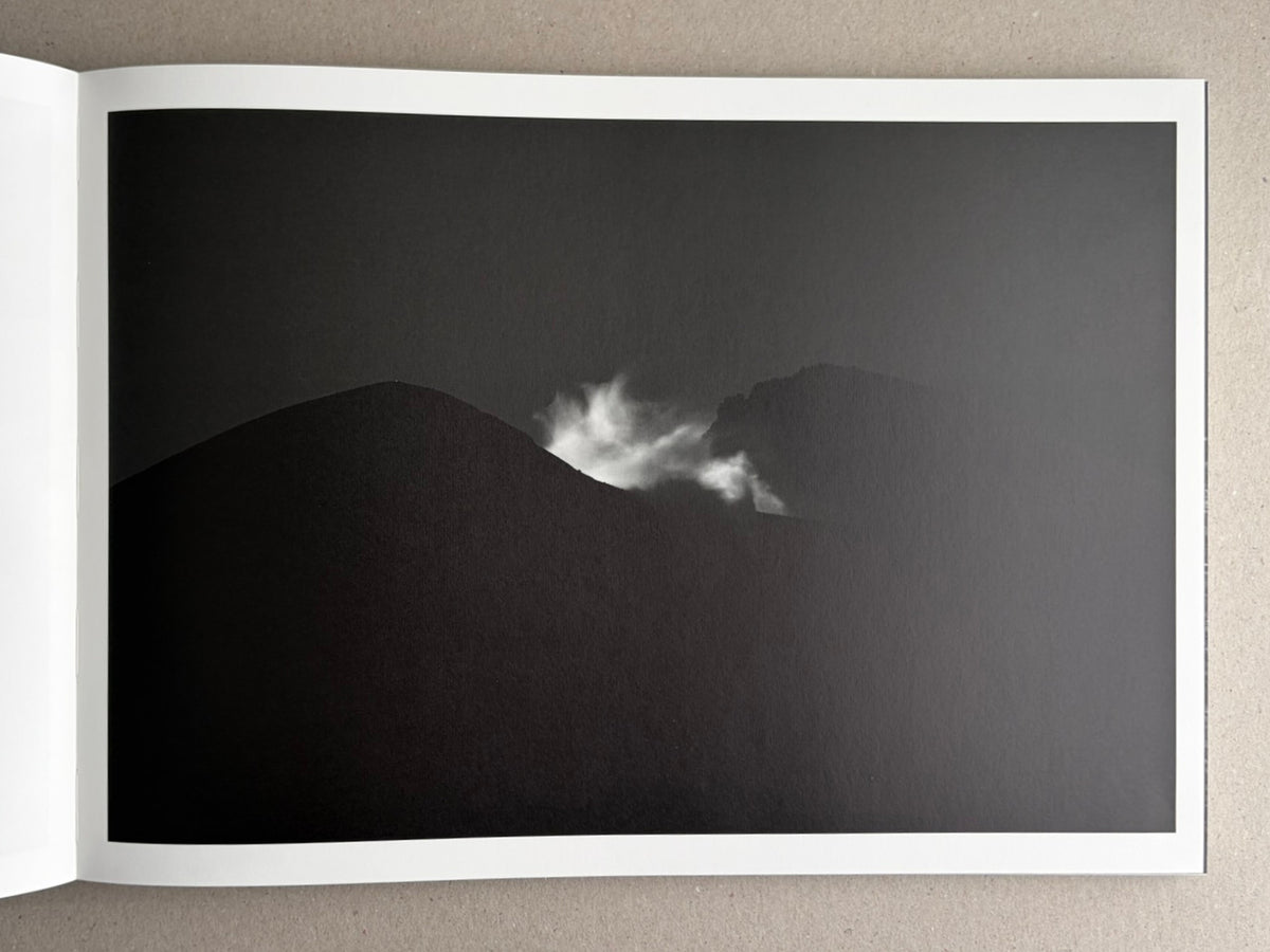 Open book spread from New Land: Pre-Hokkaido Landscapes by Toshiki Nakanishi, showing a dark landscape photograph with a bright plume of steam or smoke between two hills.