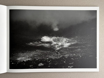 Open photobook page showing a black-and-white landscape photograph of a dark, misty rocky scene.