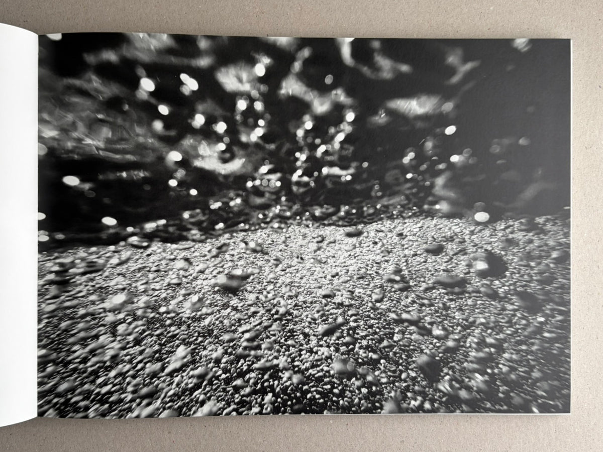 Open photobook spread from New Land: Pre-Hokkaido Landscapes by Toshiki Nakanishi, showing a black-and-white abstract landscape image with blurred light reflections and a textured surface.