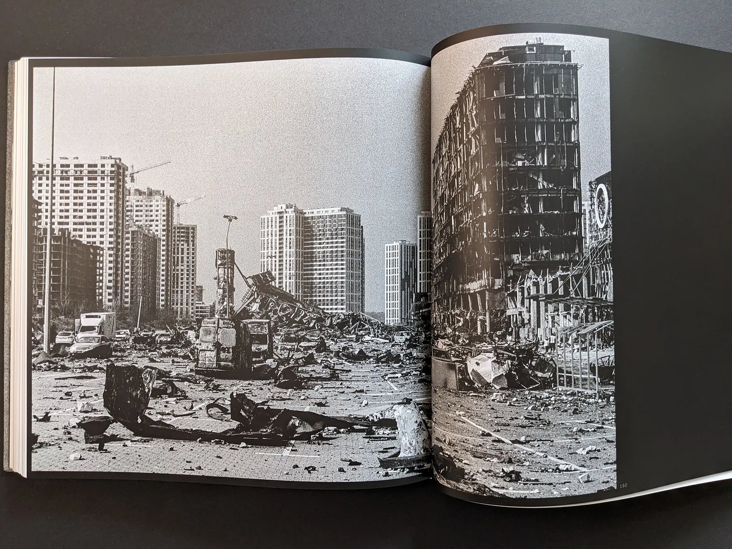 Oleksandr Glyadyelov war photograph of a devastated Ukrainian city with burned vehicles and a ruined office building among high-rise blocks.