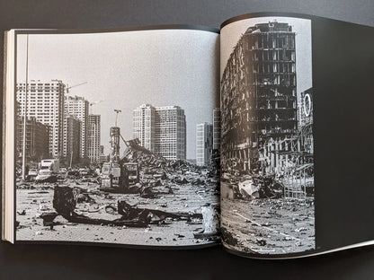 Oleksandr Glyadyelov war photograph of a devastated Ukrainian city with burned vehicles and a ruined office building among high-rise blocks.