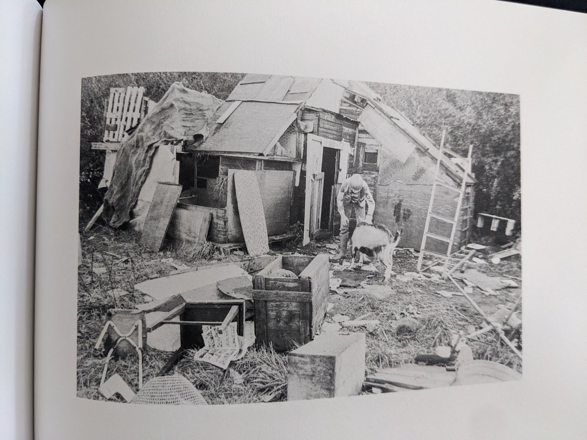 Black-and-white photograph of a child beside a goat and a makeshift hut in a cluttered outdoor setting.