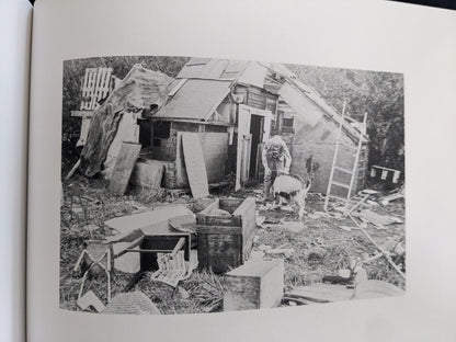 Black-and-white photograph of a child beside a goat and a makeshift hut in a cluttered outdoor setting.