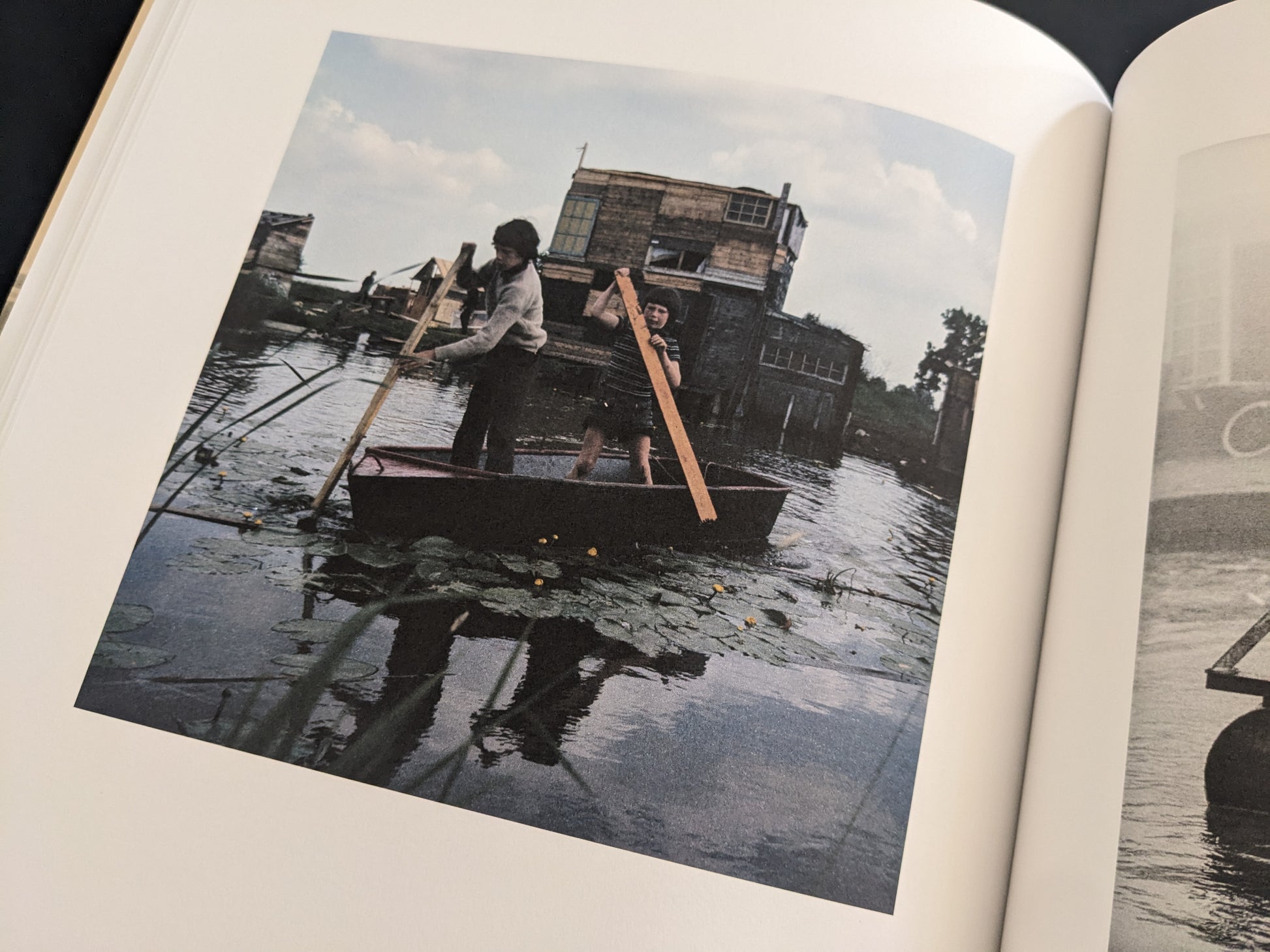 Colour photograph of two children rowing a small boat on water in front of a large wooden structure.