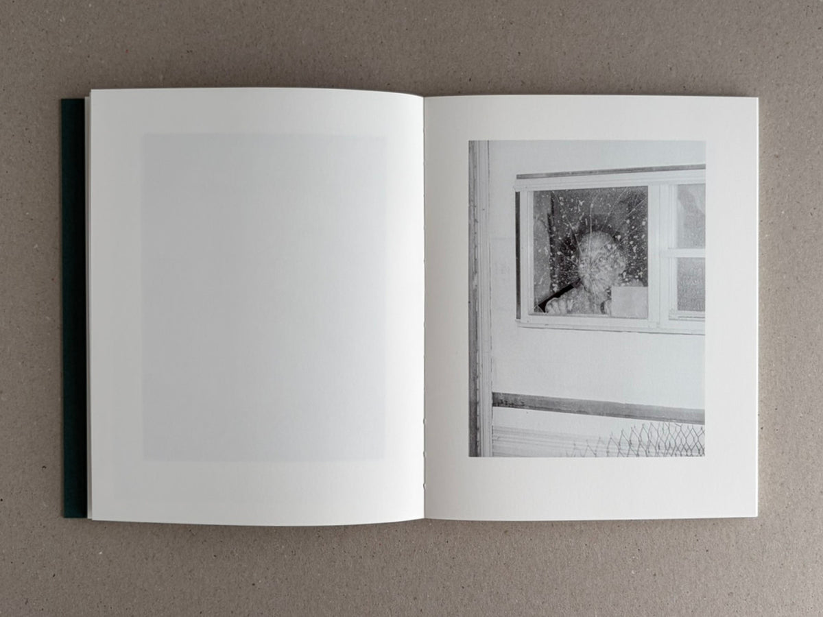 Open Sleep Creek photobook spread on a neutral surface, with a mostly blank left page and a black-and-white image on the right page showing a figure behind a window.