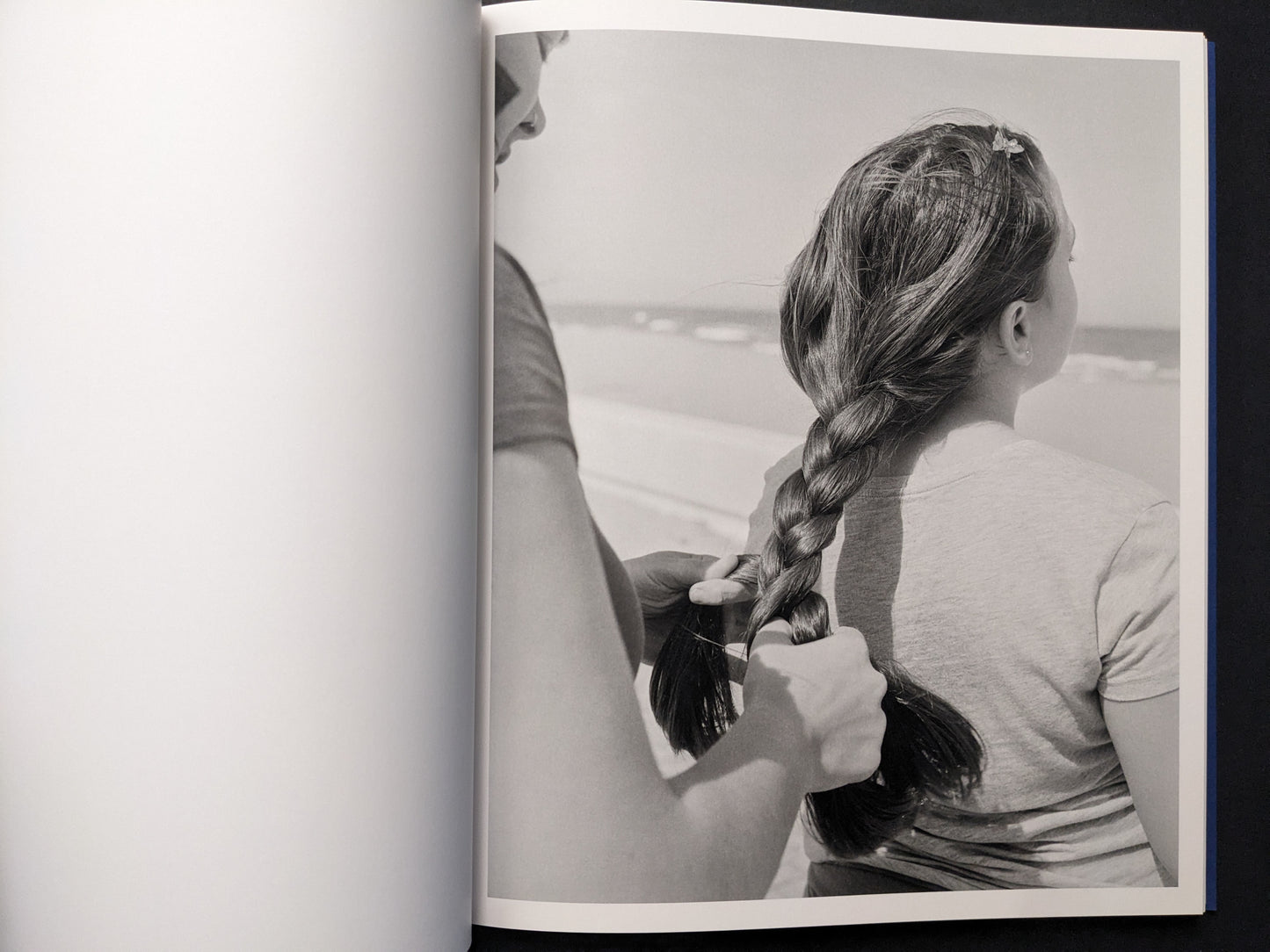 Black-and-white photograph of a person braiding a girl’s hair with the sea in the background.