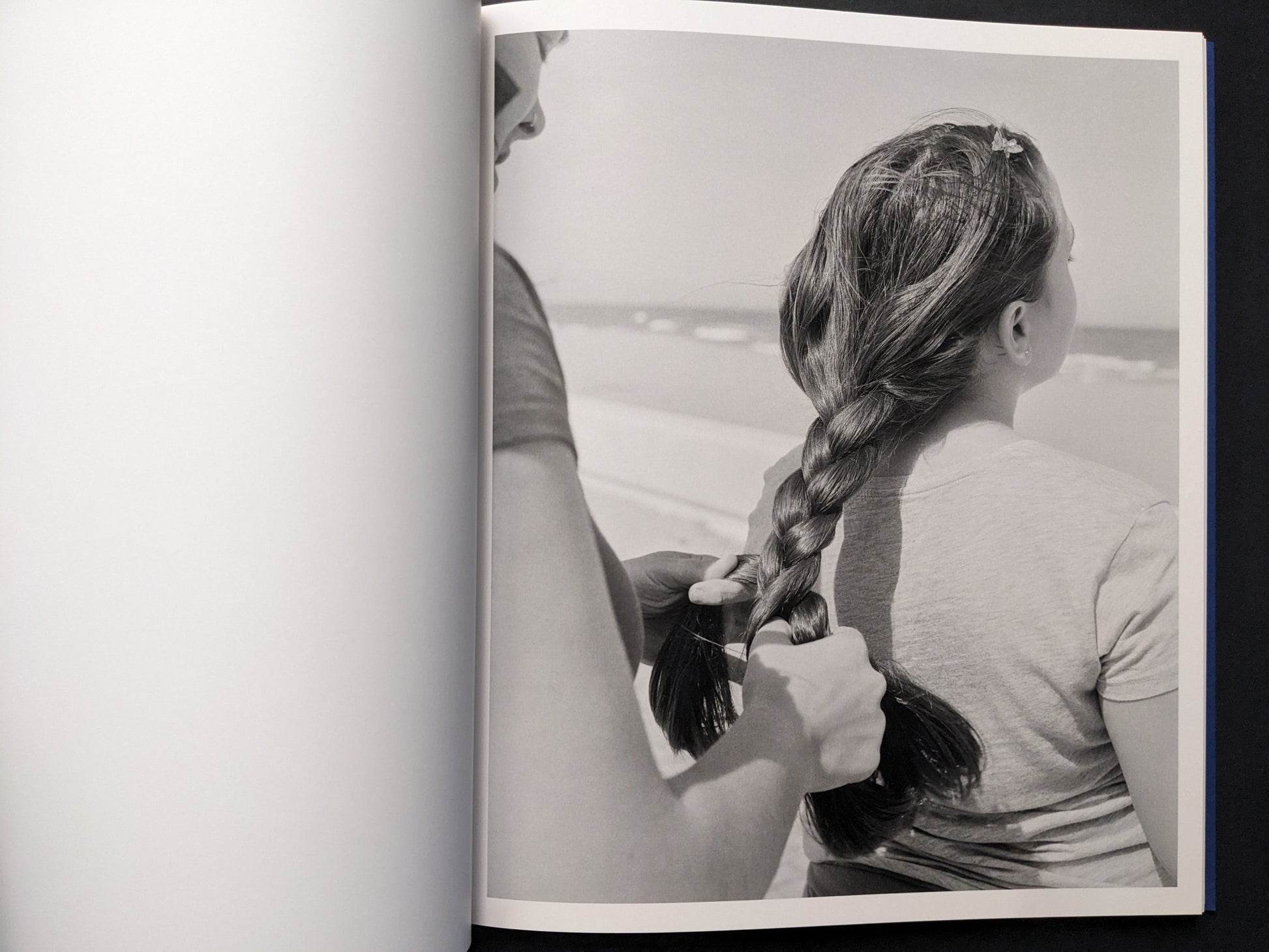 Black-and-white photograph of a person braiding a girl’s hair with the sea in the background.