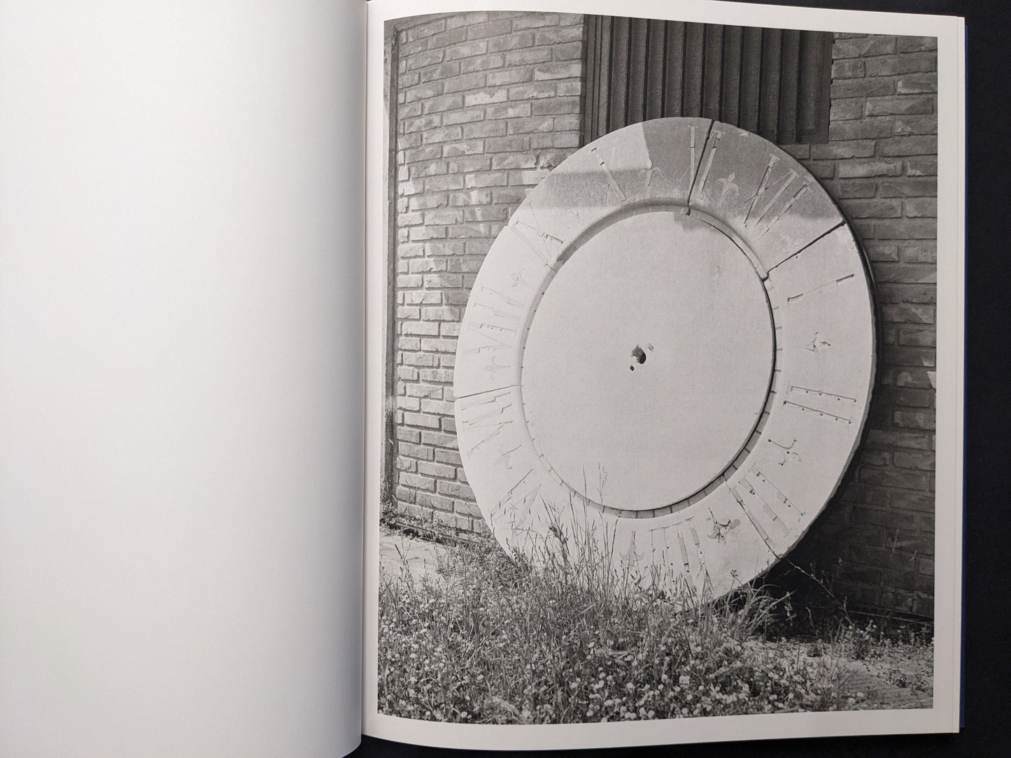 Black-and-white photograph of a large circular structure leaning against a brick wall with grass in front.
