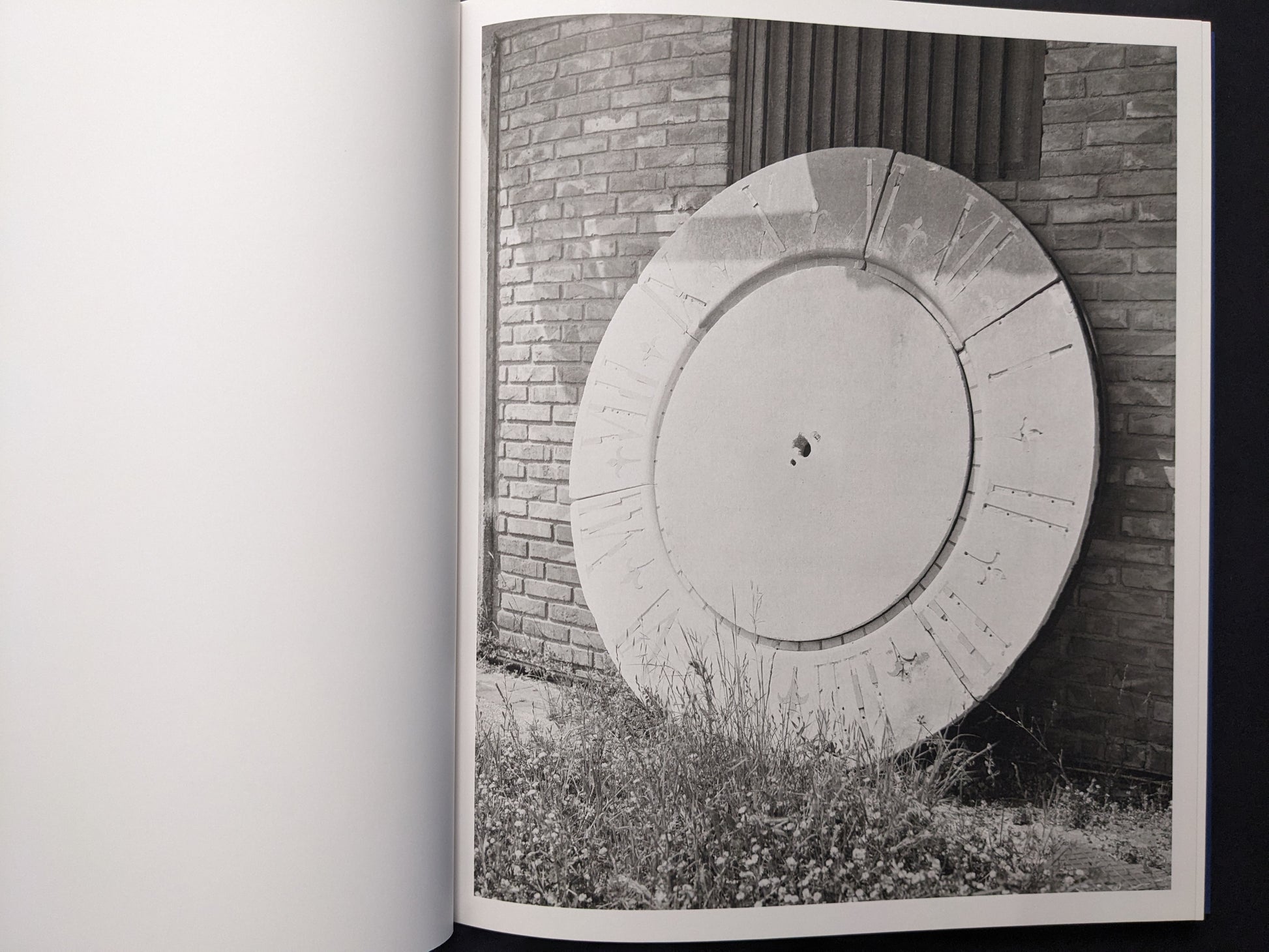 Black-and-white photograph of a large circular structure leaning against a brick wall with grass in front.