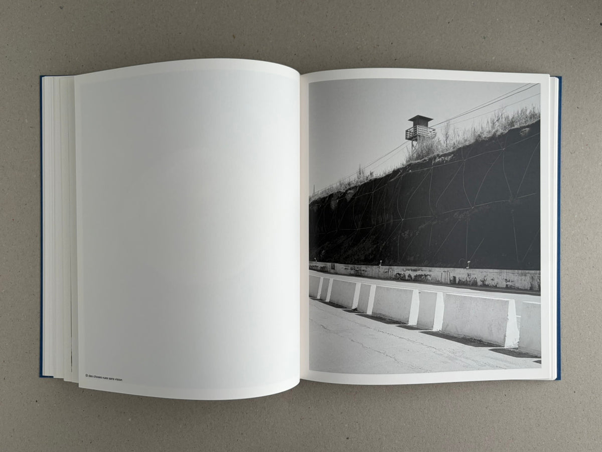 Open copy of Sub Sole by Massao Mascaro showing a black-and-white spread with a blank left page and a right-page photograph of a roadside barrier beside a steep wall and watchtower.