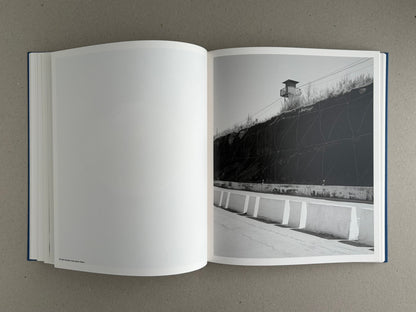 Open copy of Sub Sole by Massao Mascaro showing a black-and-white spread with a blank left page and a right-page photograph of a roadside barrier beside a steep wall and watchtower.