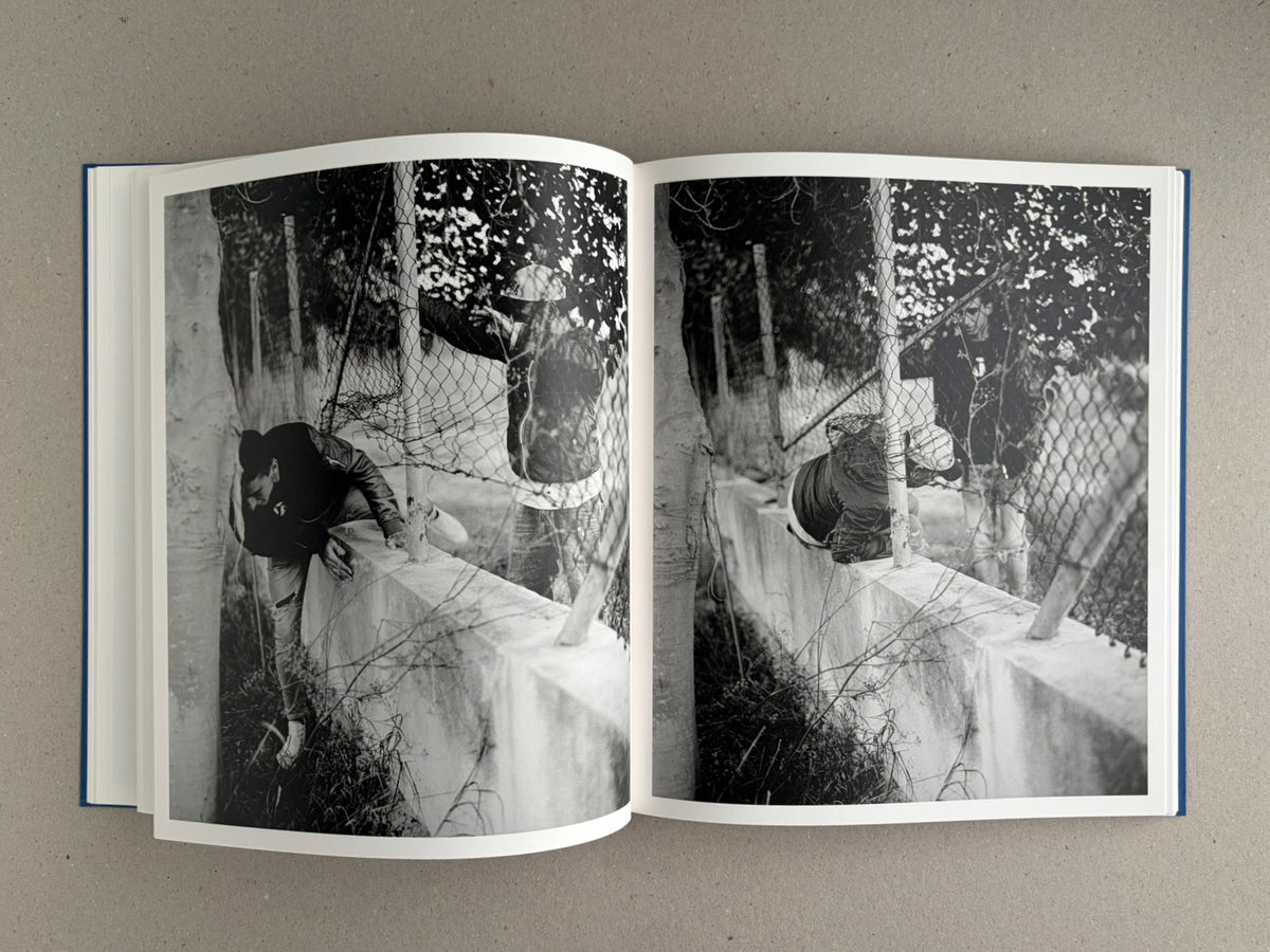 Sub Sole – Massao Mascaro open to a black-and-white two-page spread showing a figure climbing over a low wall behind wire fencing.