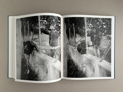 Sub Sole – Massao Mascaro open to a black-and-white two-page spread showing a figure climbing over a low wall behind wire fencing.
