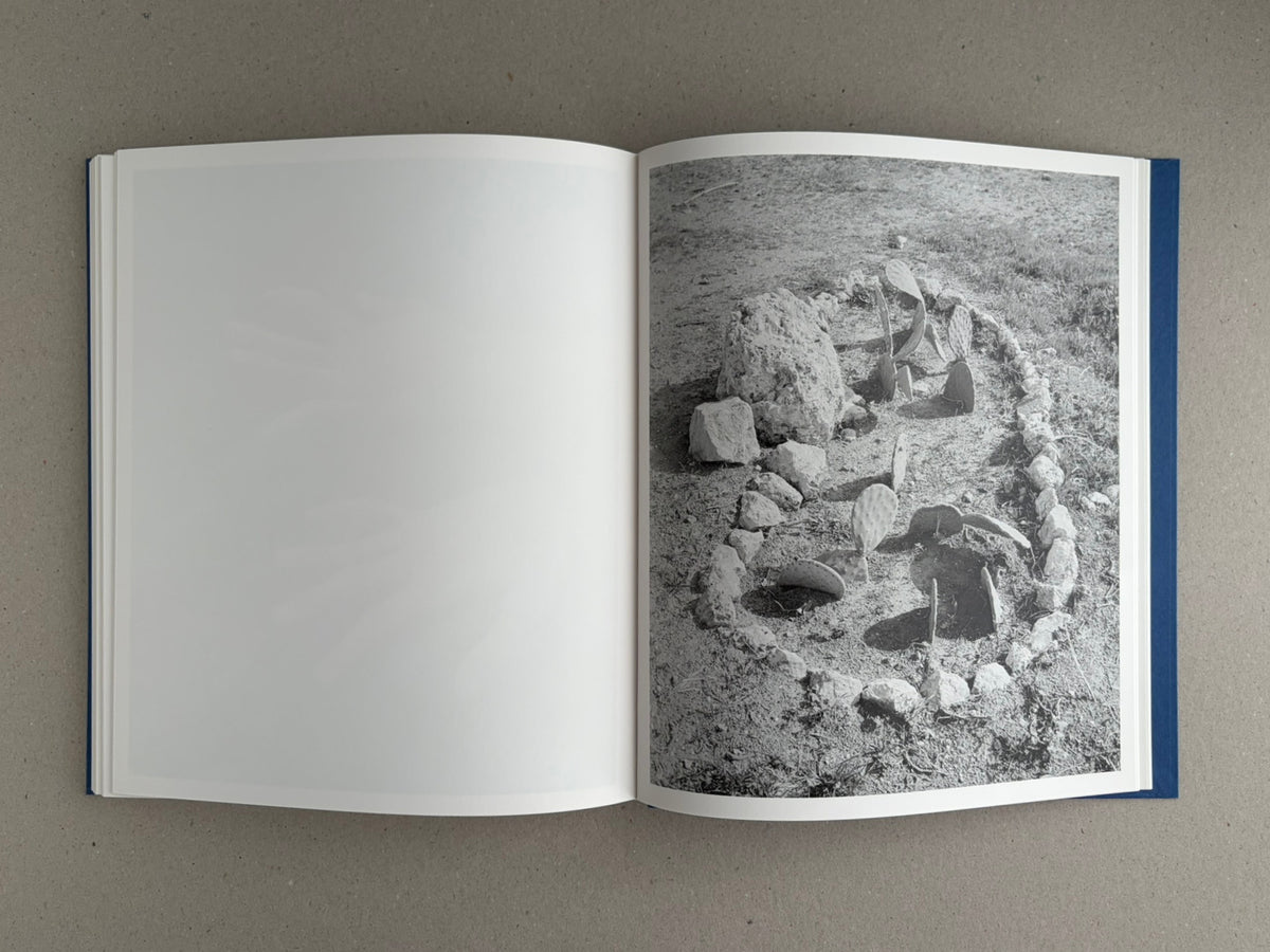 Open book spread from Sub Sole – Massao Mascaro, with a blank left page and a black-and-white photograph on the right page showing a small stone arrangement on bare ground.