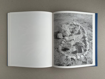 Open book spread from Sub Sole – Massao Mascaro, with a blank left page and a black-and-white photograph on the right page showing a small stone arrangement on bare ground.