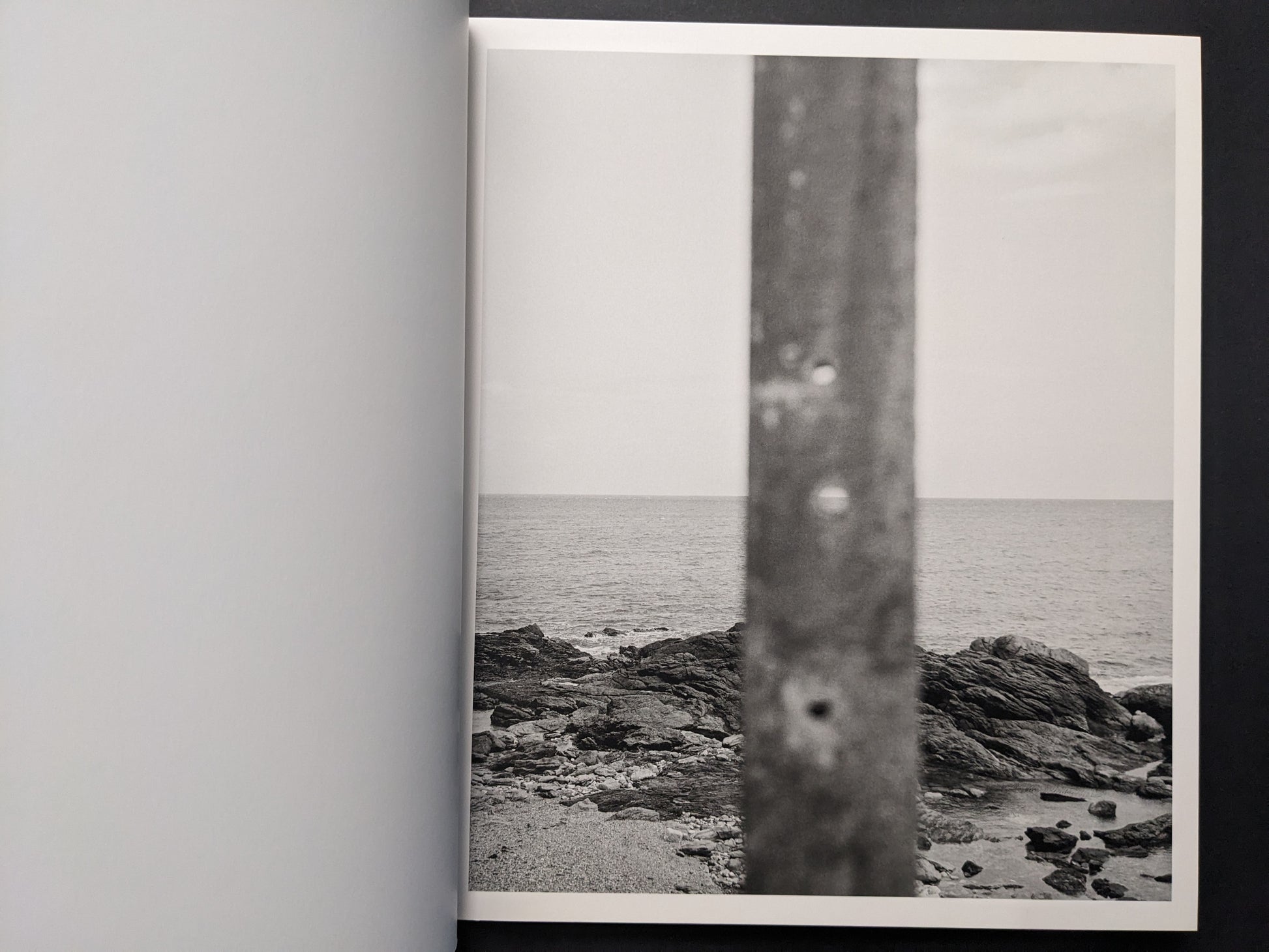 Interior photograph from Sub Sole by Massao Mascaro showing a rocky coastline and calm sea under a pale sky, partially obscured by a blurred vertical element.