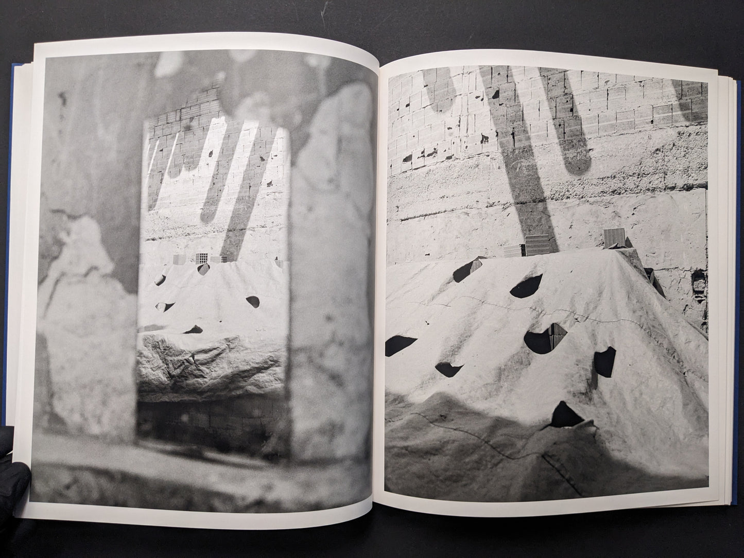 Photobook spread with a quarry-like wall and a tarp-covered structure seen through a blurred foreground frame.