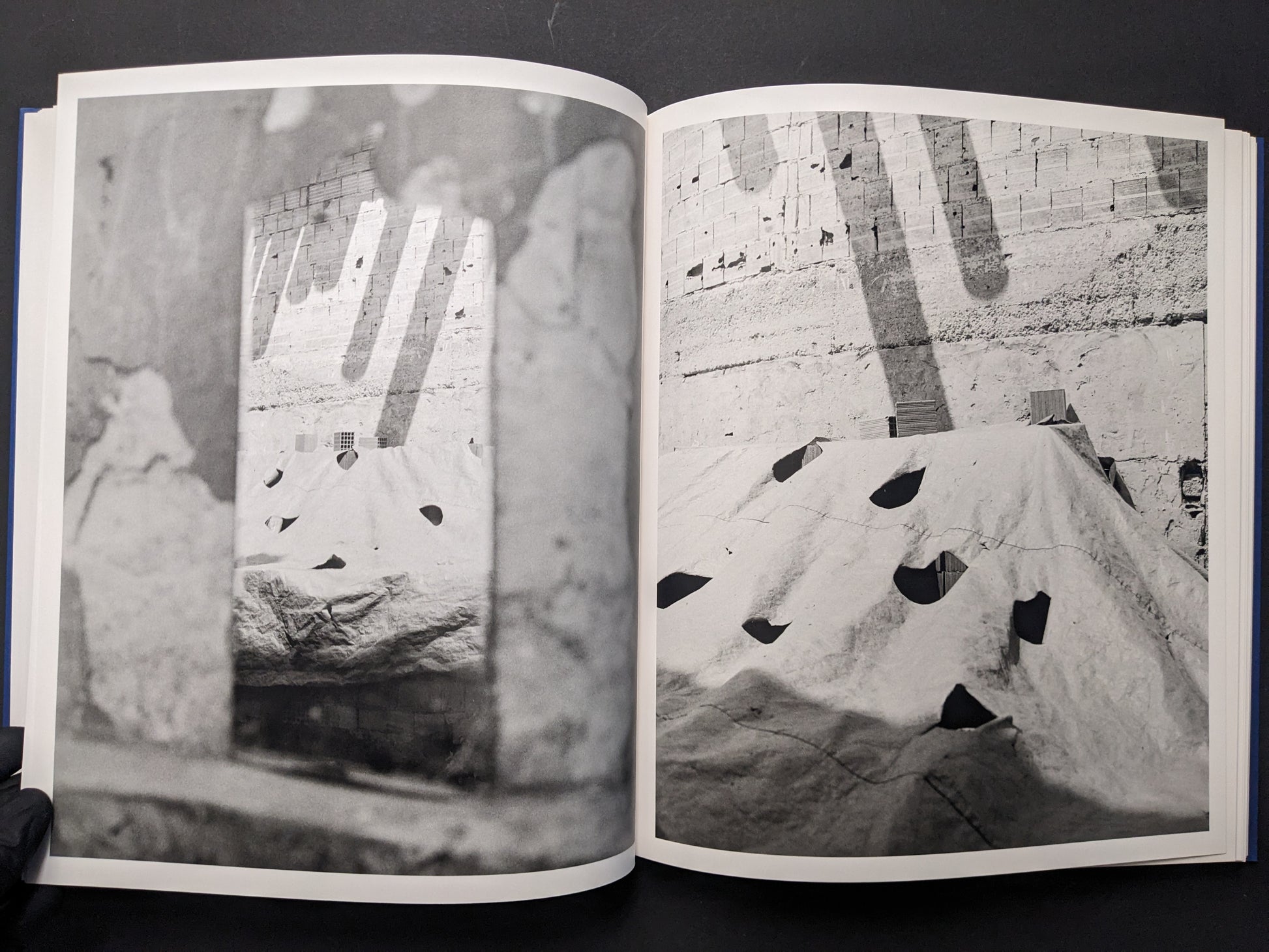 Photobook spread with a quarry-like wall and a tarp-covered structure seen through a blurred foreground frame.