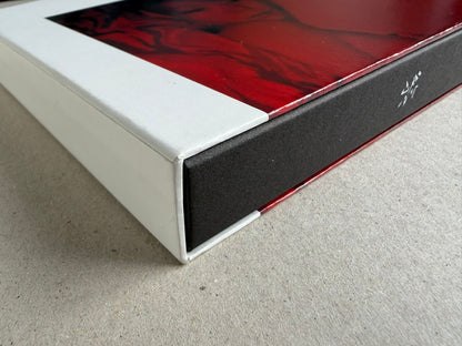 Close-up of the corner of the Surveillance – Tsutomu Yamagata book or box, showing a white outer sleeve, black side panel, and red printed cover edge.