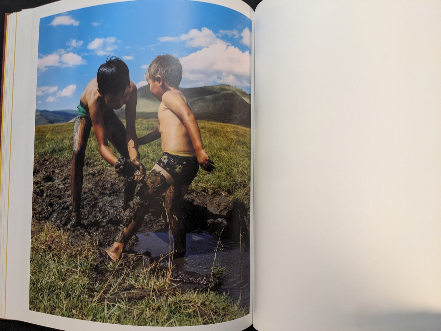 Photograph of two children playing in mud on grassland under a bright blue sky.
