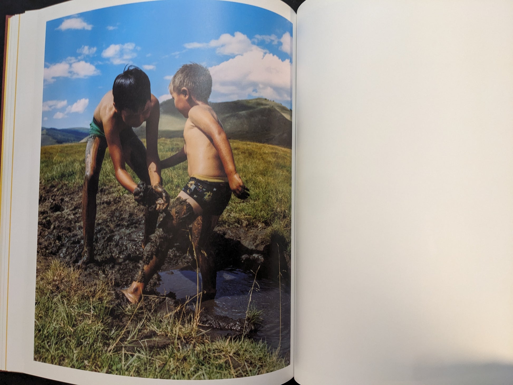 Photograph of two children playing in mud on grassland under a bright blue sky.