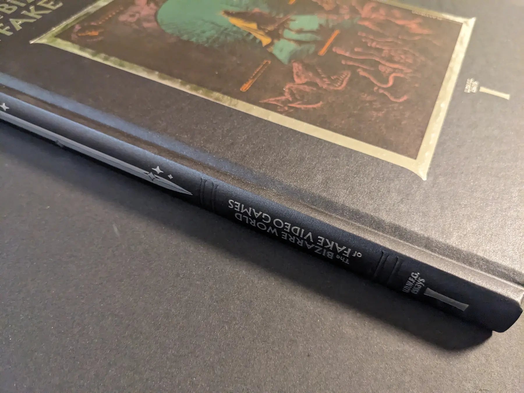 Angled close-up of the spine of The Bizarre World of Fake Video Games hardcover, featuring the title and a silver sword icon on textured black cloth.