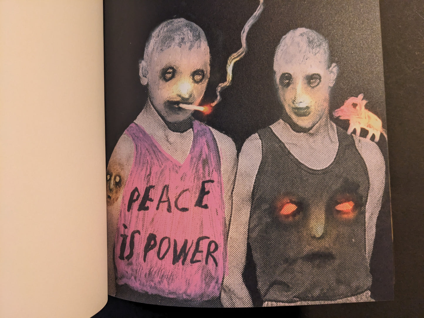 Artwork from The Fire That Drank The Shadow by Julia Soboleva showing two pale figures with dark eyes; one smokes a cigarette and wears a top with “PEACE IS POWER” text and a large face motif with glowing eyes.