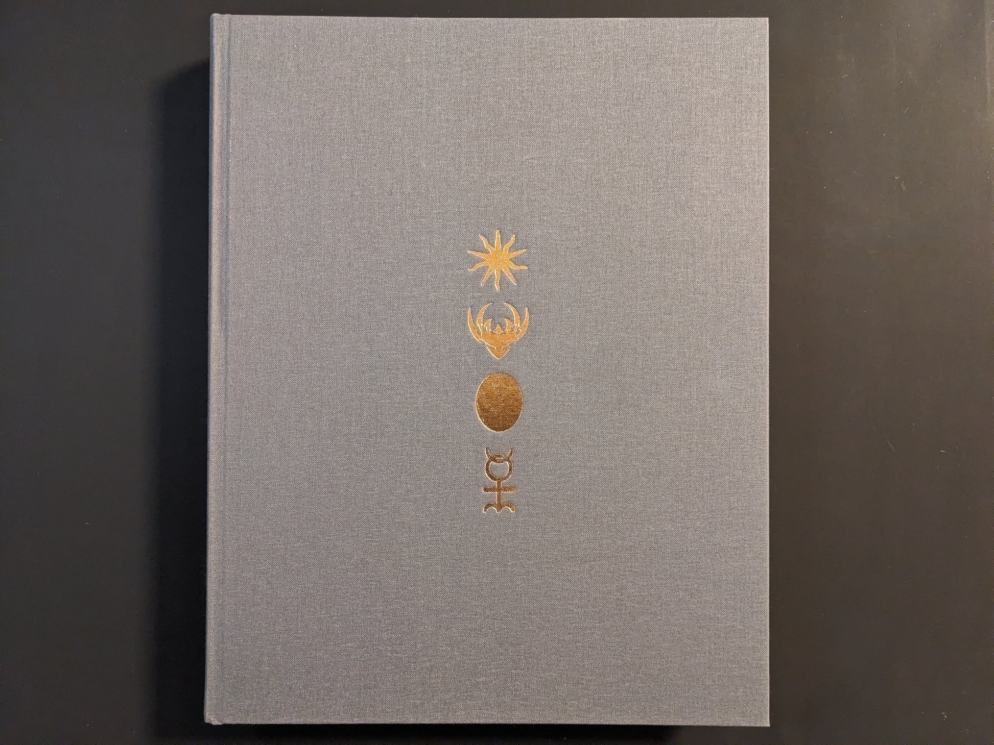 Front cover of The Universe is a Haunted House: COIL through their art & archives, gray cloth hardcover with gold embossed symbols.
