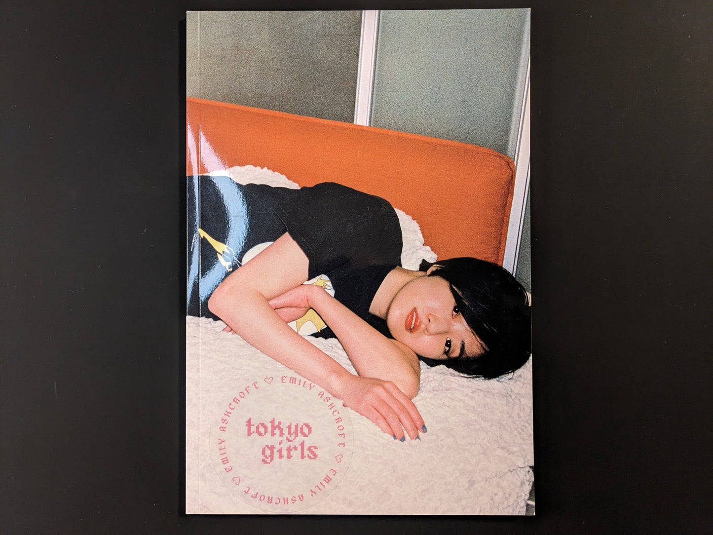 Front cover of Tokyo Girls by Emily Ashcroft, featuring a young woman lying on an orange bench, looking directly at the camera.