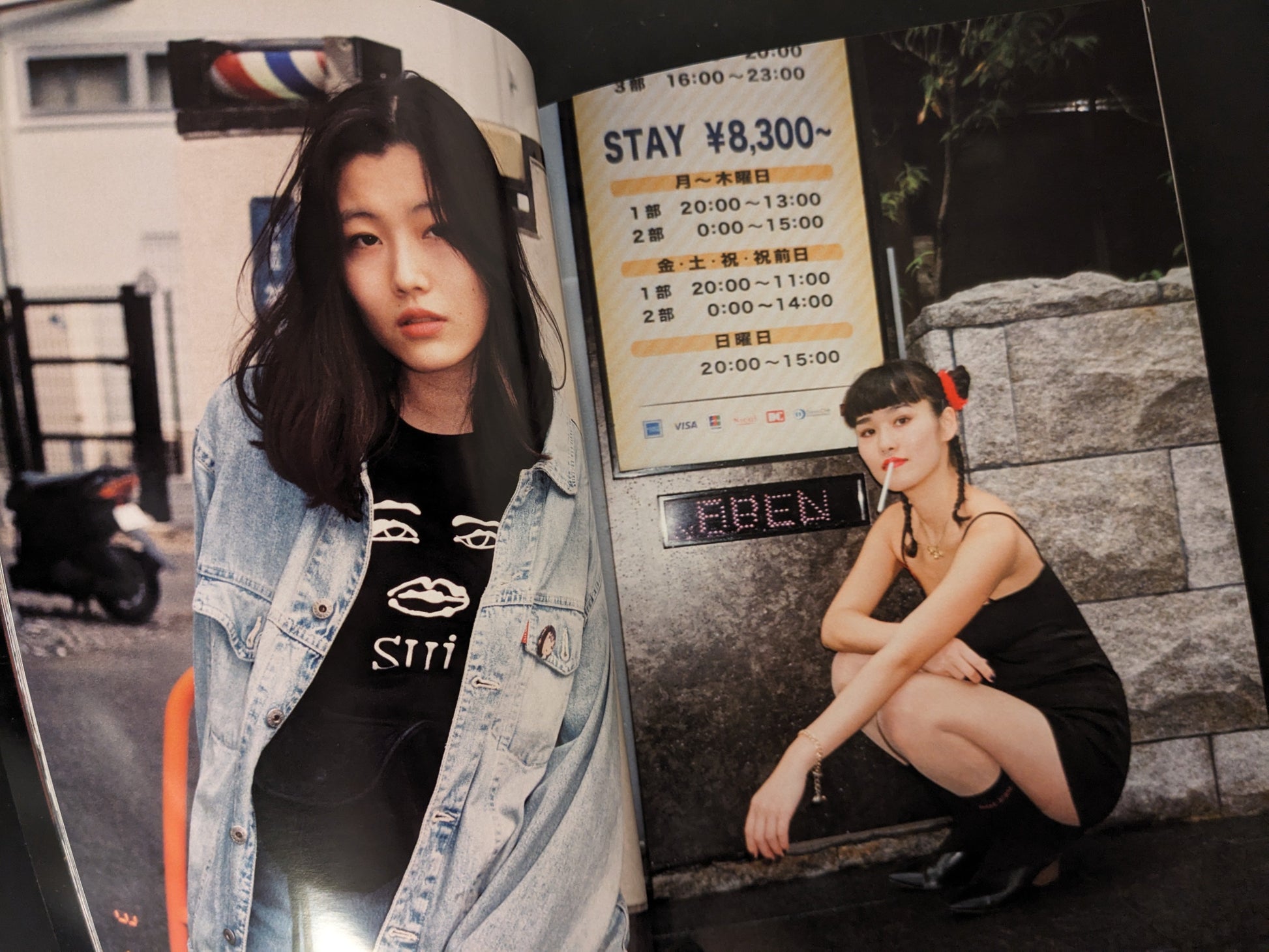 Portrait of two women, one standing against a wall in a black outfit, another crouched in front of a signboard, night street scene.