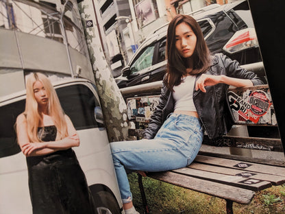 Two women in casual streetwear, one sitting on a bench with stickers, another standing confidently in the background.