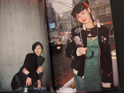 Two women at night in Tokyo, one squatting against a wall, another holding a transparent umbrella on a rainy street.