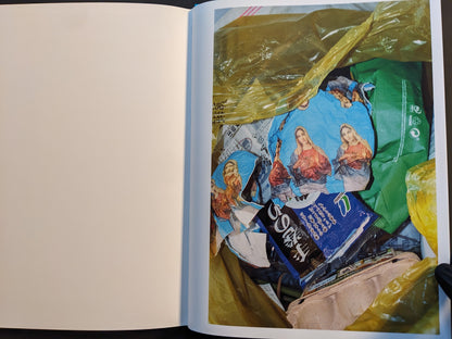 Open spread from ¥€$U$ (JESUS) showing torn Virgin Mary wrappers in a trash bag with other rubbish.