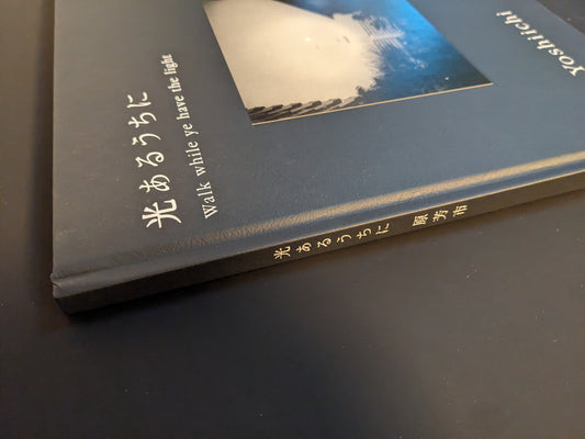 Close-up of the spine of Yoshiichi Hara’s photobook “Walk while ye have the light” with the Japanese and English title stamped in white on the grey cover.