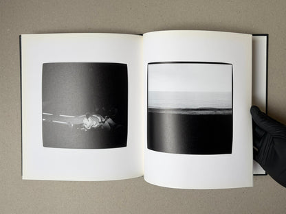 Open photobook spread from Walk while ye have the light by Yoshiichi Hara, showing two black-and-white photographs: a small group seated on a dark road at left and a seascape at right.
