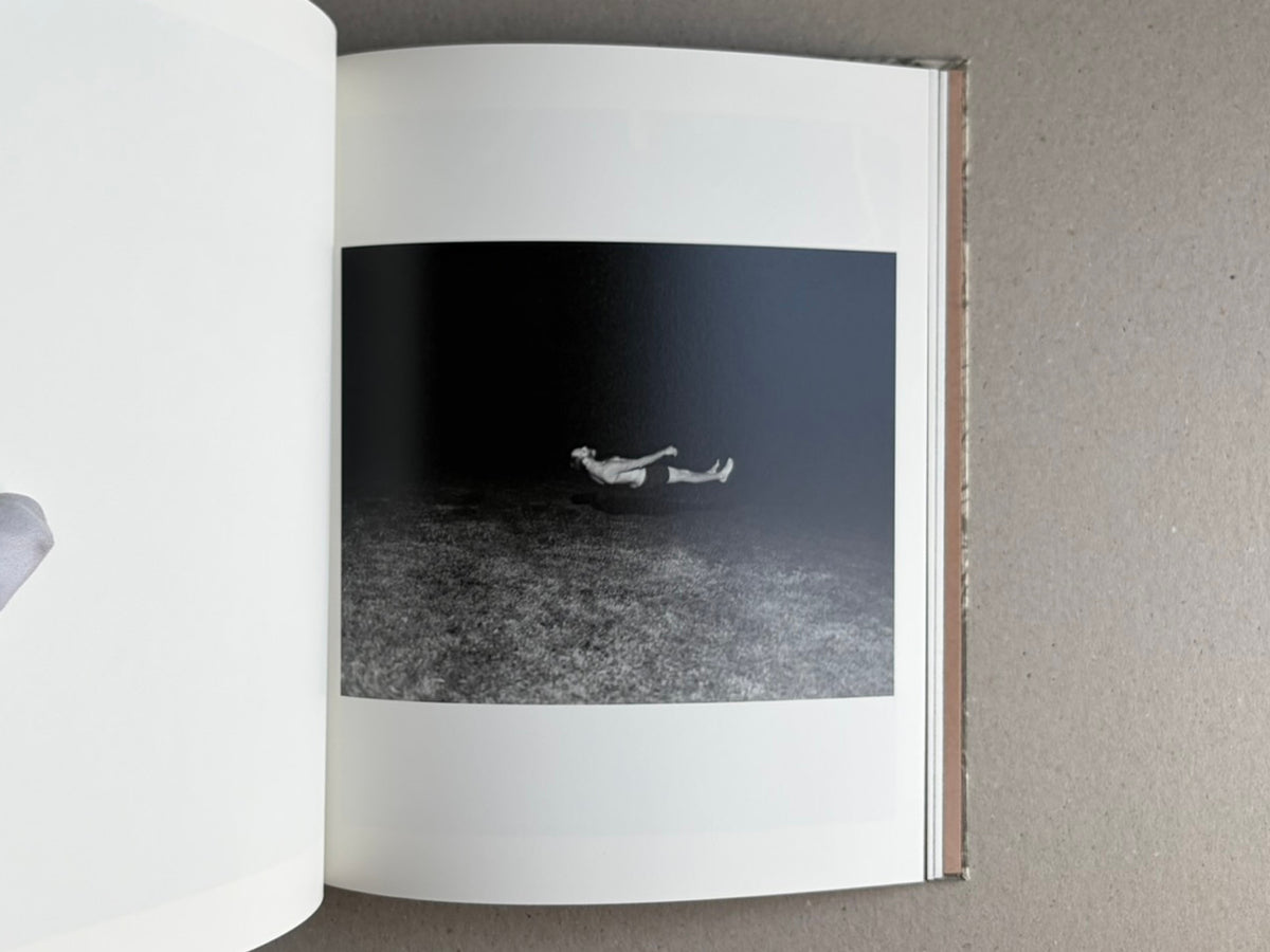 Open photobook spread from What the Rain Might Bring by Dylan Hausthor, showing a black-and-white image of a figure floating or lying horizontally in a dark landscape.