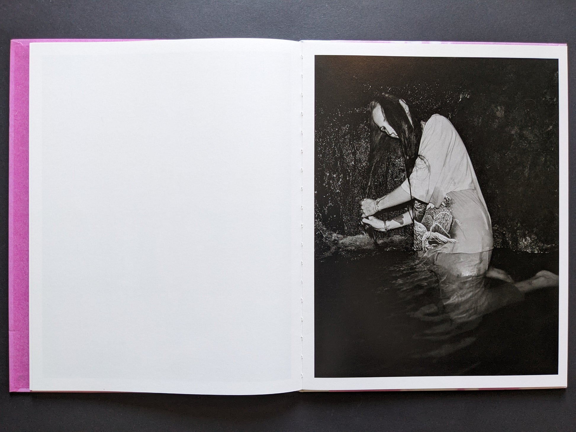 Open spread with a blank left page and a right-hand portrait of a woman kneeling in water and washing her hair in a cave-like setting.