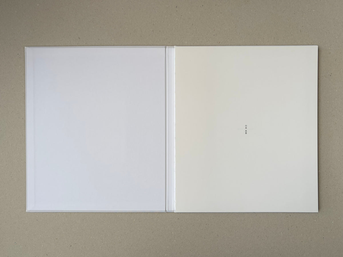 Open Yamamoto Masao photobook showing blank white endpapers and a small vertical Japanese title mark on the right page.